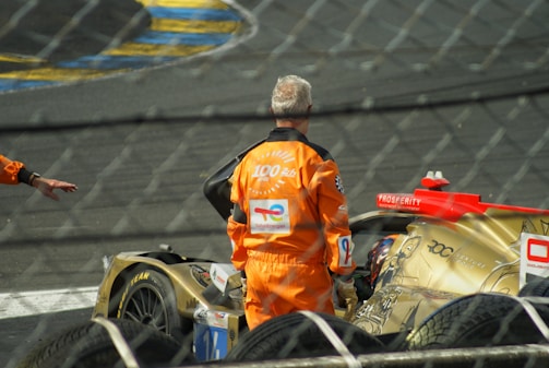A race car with a golden and red design is positioned on a track, with two individuals in orange uniforms nearby. The setting suggests a motorsport event, possibly during a pit stop or post-race scenario. Protective fencing is visible in the foreground.