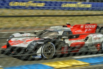 A high-speed racing car is captured in motion on a race track. The car features prominent branding and sponsorship logos, including red, white, and grey color schemes. An out-of-focus background suggests high velocity, and the image is partially obscured by a safety fence.