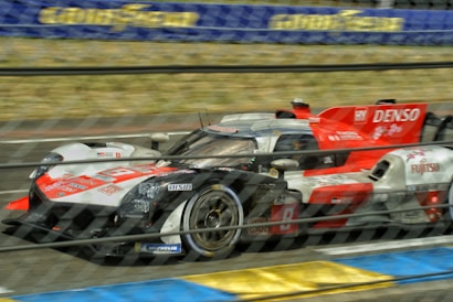 A high-speed racing car is captured in motion on a race track. The car features prominent branding and sponsorship logos, including red, white, and grey color schemes. An out-of-focus background suggests high velocity, and the image is partially obscured by a safety fence.