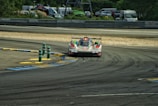 A race car is navigating through a track with painted curbs in blue and yellow. The car is designed for high-speed racing, featuring a sleek, low-profile design. The environment includes barriers and a backdrop of parked vehicles and green foliage.