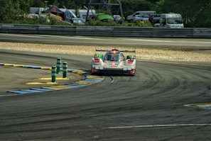 A race car is navigating through a track with painted curbs in blue and yellow. The car is designed for high-speed racing, featuring a sleek, low-profile design. The environment includes barriers and a backdrop of parked vehicles and green foliage.