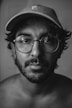 The image features a portrait of a person with a beard and large round glasses, wearing a cap. The lighting creates a high contrast black and white effect, highlighting facial features and textures.