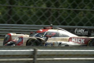 A high-speed race car is captured in motion on a racetrack. The car features prominent branding and sponsor logos, and is surrounded by a metallic fence barrier. The track background is blurred, emphasizing the speed of the vehicle.