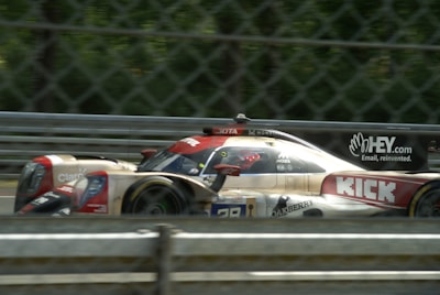A high-speed race car is captured in motion on a racetrack. The car features prominent branding and sponsor logos, and is surrounded by a metallic fence barrier. The track background is blurred, emphasizing the speed of the vehicle.