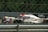 A high-speed race car is captured in motion on a racetrack. The car features prominent branding and sponsor logos, and is surrounded by a metallic fence barrier. The track background is blurred, emphasizing the speed of the vehicle.