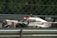 A high-speed race car is captured in motion on a racetrack. The car features prominent branding and sponsor logos, and is surrounded by a metallic fence barrier. The track background is blurred, emphasizing the speed of the vehicle.