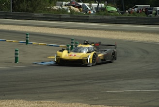 A racing car speeds along a track, taking a sharp turn. The car has a sleek design with a prominent yellow and black color scheme. The environment includes barriers with colored markers and a backdrop of trees, tents, and spectators in the distance.