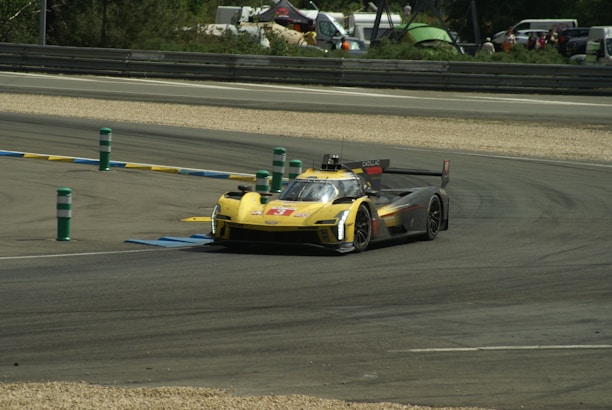 A racing car speeds along a track, taking a sharp turn. The car has a sleek design with a prominent yellow and black color scheme. The environment includes barriers with colored markers and a backdrop of trees, tents, and spectators in the distance.