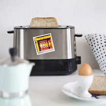 A stainless steel toaster with a slice of bread popping out, adorned with a colorful sticker that reads 'HELLO.' The setting includes a polka dot pillow, a small coffee maker, and a breakfast setup with a boiled egg in an egg cup and slices of bread on a plate.