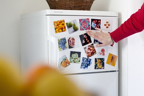 A hand with a red sleeve is touching the door of a white refrigerator covered with colorful pictures of various fruits such as oranges, pears, blueberries, pomegranates, and kiwis. On top of the refrigerator is a woven basket.