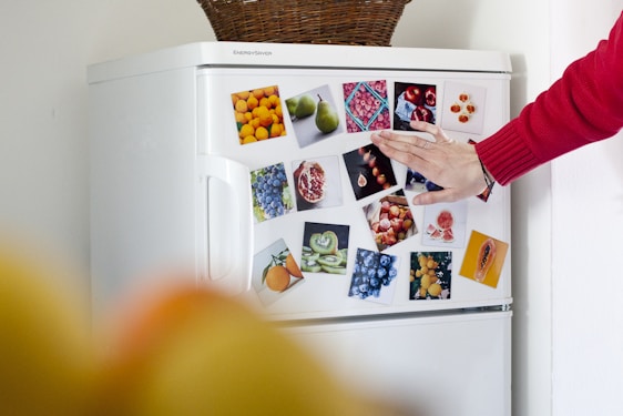 A hand with a red sleeve is touching the door of a white refrigerator covered with colorful pictures of various fruits such as oranges, pears, blueberries, pomegranates, and kiwis. On top of the refrigerator is a woven basket.