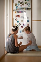 Two children are sitting on the floor in front of a refrigerator covered in colorful magnets and photographs. The refrigerator door is decorated with various letter and number magnets, as well as small images and stickers. The children appear to be engaged in a conversation or activity involving the magnets.