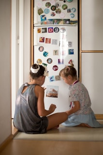Two children are sitting on the floor in front of a refrigerator covered in colorful magnets and photographs. The refrigerator door is decorated with various letter and number magnets, as well as small images and stickers. The children appear to be engaged in a conversation or activity involving the magnets.