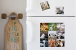 A white refrigerator decorated with multiple animal-themed photographs. The top section features two smaller images of a dog in a forest and a tabby cat. The lower section displays a collage of several animals including ducklings, a rabbit, geese, turkeys, cows, pigs, a goat, a chicken, and a horse. To the left of the fridge, a longboard with stylized text is leaning against the wall.