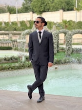 A man in a black suit and sunglasses stands confidently by a pool in a landscaped garden. The background features greenery, decorative arches adorned with flowers, and a stone wall.