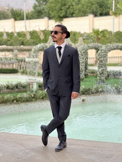 A man in a black suit and sunglasses stands confidently by a pool in a landscaped garden. The background features greenery, decorative arches adorned with flowers, and a stone wall.