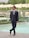 A man in a black suit and sunglasses stands confidently by a pool in a landscaped garden. The background features greenery, decorative arches adorned with flowers, and a stone wall.