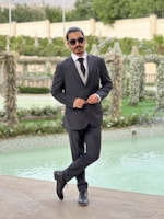 A man in a formal suit stands with one leg crossed over the other, wearing sunglasses and posing confidently beside a decorative pool. The background features lush greenery and intricate garden structures.