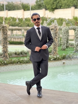 A man in a formal suit stands with one leg crossed over the other, wearing sunglasses and posing confidently beside a decorative pool. The background features lush greenery and intricate garden structures.