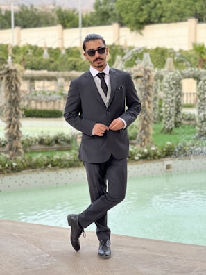 A man in a formal suit stands with one leg crossed over the other, wearing sunglasses and posing confidently beside a decorative pool. The background features lush greenery and intricate garden structures.