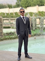 A man in a dark suit and tie stands confidently by a pool, wearing sunglasses and posing in a well-manicured garden. The background features greenery and elegantly arranged plants, creating a sophisticated outdoor setting.