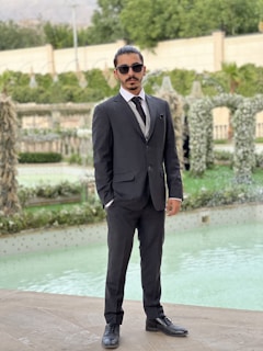 A man in a dark suit and tie stands confidently by a pool, wearing sunglasses and posing in a well-manicured garden. The background features greenery and elegantly arranged plants, creating a sophisticated outdoor setting.