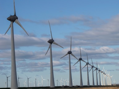 A row of large wind turbines stretches into the distance over a flat landscape. The blades of the turbines are pointed in different directions, indicating varying wind directions. Above, fluffy clouds are scattered across a blue sky.