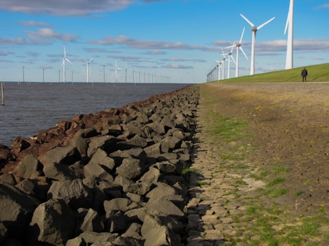 A coastal landscape featuring a row of wind turbines extending into the distance. The foreground includes a rocky embankment leading to grassy terrain, with a solitary person walking along a path. The sky is partly cloudy and the sea appears calm.
