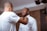 Two men are in a room, with one helping the other adjust his bow tie. One man is wearing a white dress shirt, while the other is in a casual white t-shirt. The setting appears personal and relaxed.