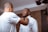 Two men are in a room, with one helping the other adjust his bow tie. One man is wearing a white dress shirt, while the other is in a casual white t-shirt. The setting appears personal and relaxed.