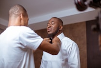 Two men are in a room, with one helping the other adjust his bow tie. One man is wearing a white dress shirt, while the other is in a casual white t-shirt. The setting appears personal and relaxed.