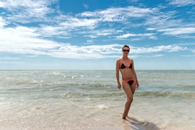 A woman wearing a black bikini with orange details stands on the beach with waves gently lapping at her feet. She is wearing sunglasses and the sky is predominantly clear with some scattered clouds. The sea water appears calm and stretches out to the horizon.