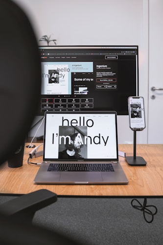 A workspace featuring a large monitor, a MacBook laptop, and a smartphone on a stand. The screens display the phrase 'Hello I'm Andy' with matching designs. The desk is wooden with minimal clutter, containing only a black cup and some cables.