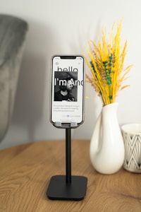 A wooden table holds a sleek black phone stand with a smartphone displaying a webpage featuring a portrait photo and introductory text in sleek typography. To the right, there's a white ceramic vase with a geometric pattern containing bright yellow and green decorative plants, contributing a pop of color. The background is soft and neutral, creating a serene atmosphere.