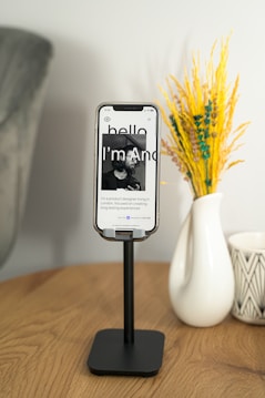 A wooden table holds a sleek black phone stand with a smartphone displaying a webpage featuring a portrait photo and introductory text in sleek typography. To the right, there's a white ceramic vase with a geometric pattern containing bright yellow and green decorative plants, contributing a pop of color. The background is soft and neutral, creating a serene atmosphere.