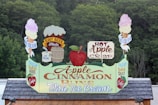 A sign prominently displaying promotions for Apple Cinnamon Buns with ice cream. It features vibrant graphics, including an apple, ice cream cones, a barrel, and the words 'Hot Apple Crisp,' 'Root Beer Floats,' and 'Fresh Baked Daily.' The backdrop consists of dense green foliage.