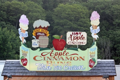 A sign prominently displaying promotions for Apple Cinnamon Buns with ice cream. It features vibrant graphics, including an apple, ice cream cones, a barrel, and the words 'Hot Apple Crisp,' 'Root Beer Floats,' and 'Fresh Baked Daily.' The backdrop consists of dense green foliage.
