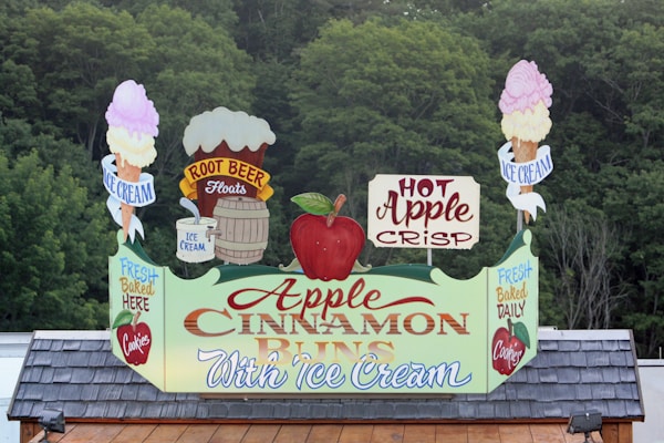 A sign prominently displaying promotions for Apple Cinnamon Buns with ice cream. It features vibrant graphics, including an apple, ice cream cones, a barrel, and the words 'Hot Apple Crisp,' 'Root Beer Floats,' and 'Fresh Baked Daily.' The backdrop consists of dense green foliage.