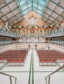 An indoor hall with a high glass ceiling and exposed iron beams. Rows of red cushioned chairs are arranged neatly, facing a central stage. The walls are adorned with a mix of brick and tiling, and green and white triangular banners hang overhead. A decorative stained glass window is prominent at the far end.