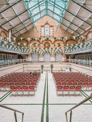 An indoor hall with a high glass ceiling and exposed iron beams. Rows of red cushioned chairs are arranged neatly, facing a central stage. The walls are adorned with a mix of brick and tiling, and green and white triangular banners hang overhead. A decorative stained glass window is prominent at the far end.