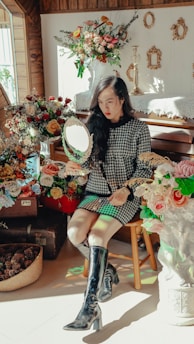 A woman with long hair wearing a checkered dress and black boots sits among a collection of vibrant floral arrangements. She holds a small hand mirror, reflecting light. The room is adorned with vintage decor including picture frames, candles, and lace tablecloths.