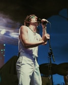 A performer is passionately singing into a microphone on stage. The person is wearing a sleeveless white shirt, white pants, and a green necklace, with distinctive sunglasses. The microphone is mounted on a stand, and the backdrop suggests an outdoor evening setting with dim lights and smoke.