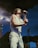 A performer is passionately singing into a microphone on stage. The person is wearing a sleeveless white shirt, white pants, and a green necklace, with distinctive sunglasses. The microphone is mounted on a stand, and the backdrop suggests an outdoor evening setting with dim lights and smoke.