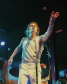A person is passionately singing on stage with one hand reaching out towards the audience. The lighting is dynamic, creating a dramatic atmosphere. The performer is wearing a white sleeveless shirt and pants, and there are musical instruments visible in the background.