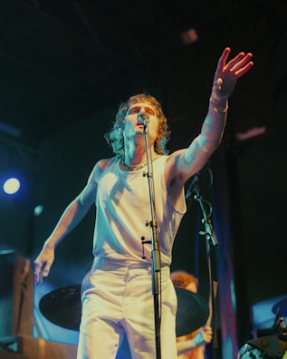 A person is passionately singing on stage with one hand reaching out towards the audience. The lighting is dynamic, creating a dramatic atmosphere. The performer is wearing a white sleeveless shirt and pants, and there are musical instruments visible in the background.
