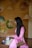 A woman in a pink traditional dress is seated on a wooden surface, holding a teapot with small tea cups and flowers nearby. Several Vietnamese hats with various decorations hang on the wall behind her.