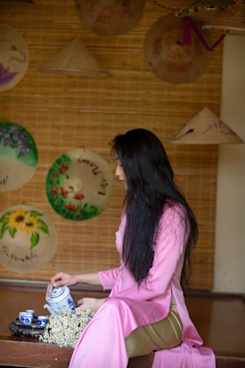 A woman in a pink traditional dress is seated on a wooden surface, holding a teapot with small tea cups and flowers nearby. Several Vietnamese hats with various decorations hang on the wall behind her.