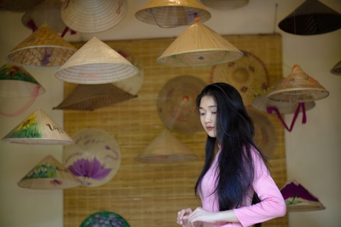 A woman in pink traditional clothing stands among various conical hats hanging from the ceiling. The hats are made of straw and some are decorated with floral patterns. The background features a beige woven texture.