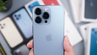 A close-up of a person holding a light blue smartphone with a triple camera setup. The phone features a prominent brand logo on its back. In the blurred background, there are several other smartphones laid out in various colors.
