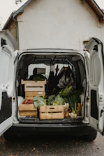 An open van parked outside a building is filled with crates of fresh vegetables, including cabbages, onions, and red bell peppers. The setup suggests an organized distribution or delivery of produce.